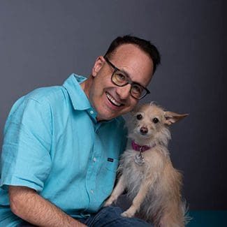 Steve Dale smiling while holding a small scruffy tan dog against a gray background.